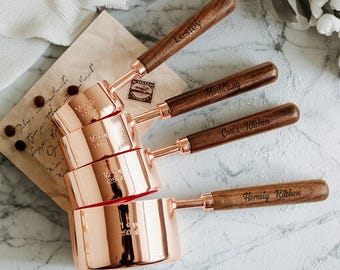May include: A set of four copper measuring cups with wooden handles. Each cup is engraved with a name or phrase: Lesley, Mindtrang, Carl's Kitchen, and Homely Kitchen. The cups are stacked, showing measurements.