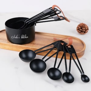 May include: A set of black measuring cups and spoons with brown leather accents. The measuring cups are nested inside each other, and the spoons are attached to a ring. The text "Julie's kitchen" is on the largest cup. The set is on a wooden tray.