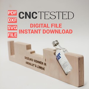 Puede incluir: Un soporte de exhibición de madera con un recorte para una botella pequeña, con el texto "DIDŽUKS VIENMĒR IR ABSOLUT'A LĪMENĪ". El soporte está etiquetado como "CNC TESTED" y "DESCARGA INSTANTÁNEA DE ARCHIVO DIGITAL". Una pequeña botella de vodka está colocada en el soporte.
