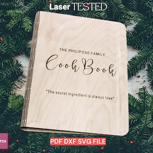 Op de afbeelding: Een houten kookboek met de tekst "THE PHILIPOSE FAMILY Cook Book" en "The secret ingredient is always love" gegraveerd op de kaft. Het kookboek staat tegen een achtergrond van groenblijvende takken en rode bessen.