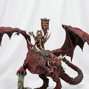 May include: A detailed miniature figure of a red dragon with large, textured wings and a rider in golden armor. The dragon has sharp teeth and claws, with a banner bearing a skull and crossbones. The figure is mounted on a stone base.
