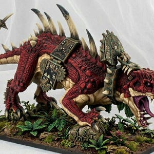 May include: A detailed miniature of a red and tan fantasy creature, possibly a dinosaur or dragon, with intricate armor and spikes. The model is posed on a base with foliage, showcasing detailed paintwork and a menacing expression.