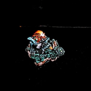May include: A miniature sculpture of a creature with an orange head, blue limbs, and green eyes, perched on a bed of green leaves and brown branches. The artwork is set against a black background, highlighting the vibrant colors and intricate details.