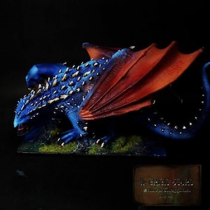 May include: A detailed miniature dragon figurine in shades of blue and red. The dragon has textured scales, sharp claws, and large wings. The wings are a vibrant red, contrasting with the blue body. The figurine is set against a dark background.