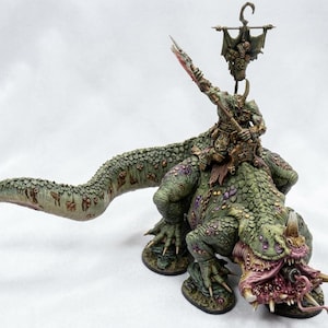 May include: A detailed miniature figure of a monstrous creature with a rider. The creature is primarily green with purple and brown accents, and has a large mouth with sharp teeth. The rider is armored and holds a weapon, with a banner above.
