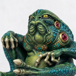 May include: A detailed figurine of a green, frog-like creature with yellow eyes and intricate teal and gold detailing. The figure has a contemplative pose, with hands resting on its legs. Red gems adorn its head and chest.
