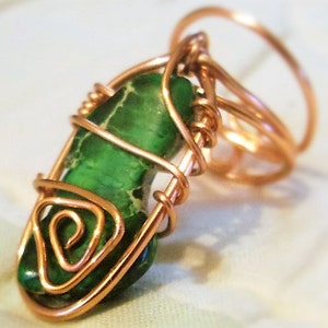 Puede incluir: Una piedra preciosa verde envuelta en alambre de cobre, creando un colgante o un anillo. El alambre está retorcido y formado en un diseño decorativo.