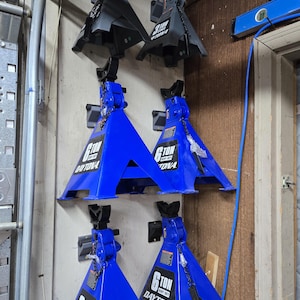 May include: A collection of automotive jack stands, some in blue and some in gray. The blue stands are labeled "6 TON DAYTONA", while the gray stands are labeled "3 TON". They are mounted on a wooden wall.