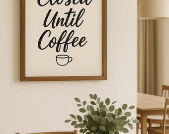 May include: A framed sign with the text "Closed Until Coffee" in black script. The sign is in a brown wooden frame and hangs on a white wall. A vase with greenery sits on a wooden table in the foreground.