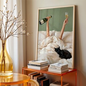 May include: A framed wall art print featuring a person in a white bed holding a champagne bottle and a glass aloft, with a black cat resting on the white bedding. The print has a sage green background and is displayed in a light wood frame.