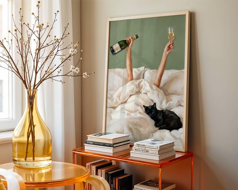 May include: A framed wall art print featuring a person in a white bed holding a champagne bottle and a glass aloft, with a black cat resting on the white bedding. The print has a sage green background and is displayed in a light wood frame.
