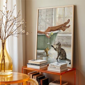 Op de afbeelding: Een ingelijst schilderij toont een persoon die ontspant in een vintage groene badkuip met een cocktail, een kat reikt naar de persoon. Een gele vaas met takken en een stapel boeken staan op de voorgrond. Het schilderij heeft een licht houten frame.