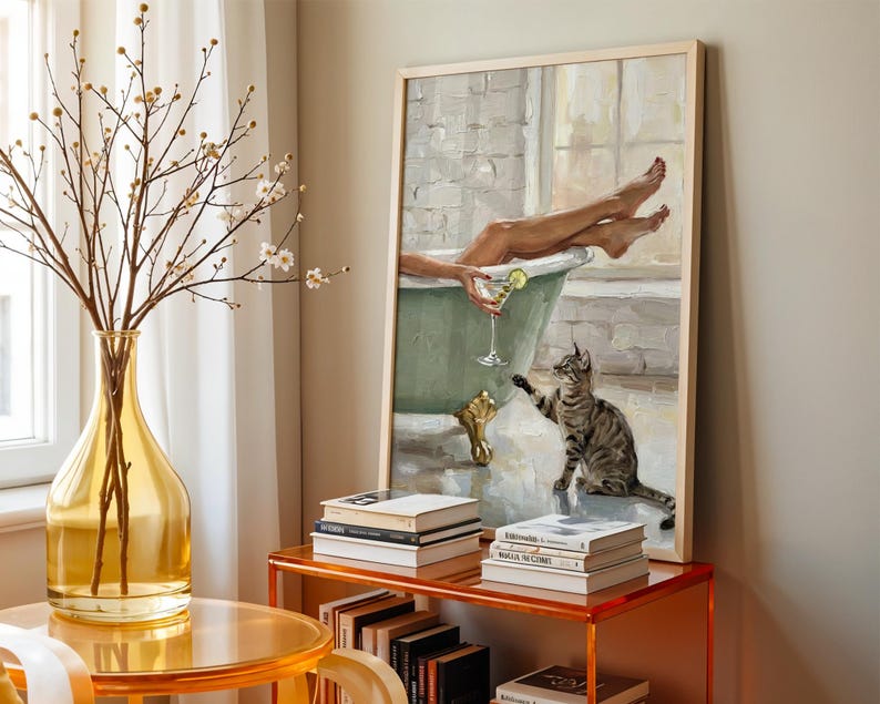 Op de afbeelding: Een ingelijst schilderij toont een persoon die ontspant in een vintage groene badkuip met een cocktail, een kat reikt naar de persoon. Een gele vaas met takken en een stapel boeken staan op de voorgrond. Het schilderij heeft een licht houten frame.