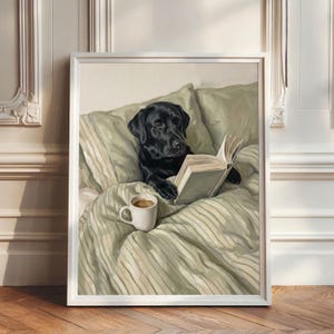 Op de afbeelding: Een ingelijste kunstdruk met een zwarte Labrador die in bed ligt met saliegroen gestreept beddengoed. De hond houdt een open boek vast met zijn poten en een witte keramische mok gevuld met koffie staat op de verfrommelde lakens.
