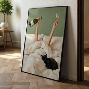 May include: A framed wall art print depicting a black cat resting on white rumpled bed sheets. Two arms extend upward from the bedding, one holding a green glass bottle and the other holding a tall, clear glass filled with a golden liquid beverage.