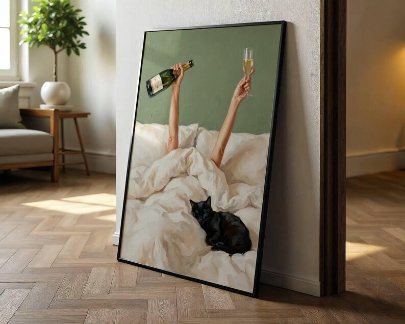 May include: A framed wall art print depicting a black cat resting on white rumpled bed sheets. Two arms extend upward from the bedding, one holding a green glass bottle and the other holding a tall, clear glass filled with a golden liquid beverage.