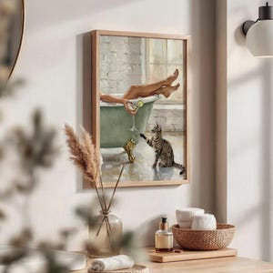 Op de afbeelding: Een ingelijst schilderij toont een persoon in een groene badkuip met een cocktail en een kat. Het kunstwerk heeft een lichte houten lijst en is aan een witte muur bevestigd. Het schilderij heeft een vintage, ontspannen thema.