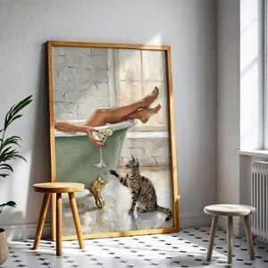 Op de afbeelding: Een ingelijst schilderij toont een ontspannende sc&egrave;ne in een badkamer. Een persoon zit in een groene badkuip en houdt een cocktail vast. Een gestreepte kat reikt naar het drankje. Het schilderij staat in een kamer met een plant en twee houten krukken.
