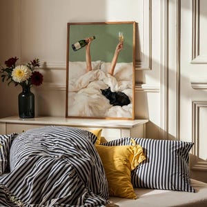May include: A framed wall art print featuring a green background with two arms raised from under white bedding, holding a champagne bottle and a glass, with a black cat resting on the bed. The print is displayed on a white wooden console table.