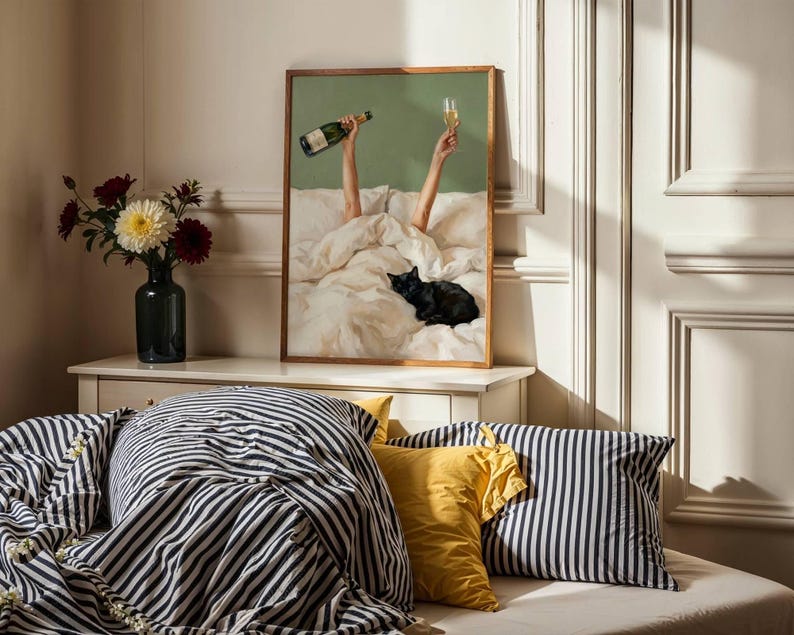 May include: A framed wall art print featuring a green background with two arms raised from under white bedding, holding a champagne bottle and a glass, with a black cat resting on the bed. The print is displayed on a white wooden console table.