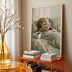 May include: A framed wall art print depicting a golden-brown curly-haired dog lying in a sage green bed, reading an open book with a white mug of coffee nearby. The artwork is displayed on a shelf alongside stacks of books and a glass vase.