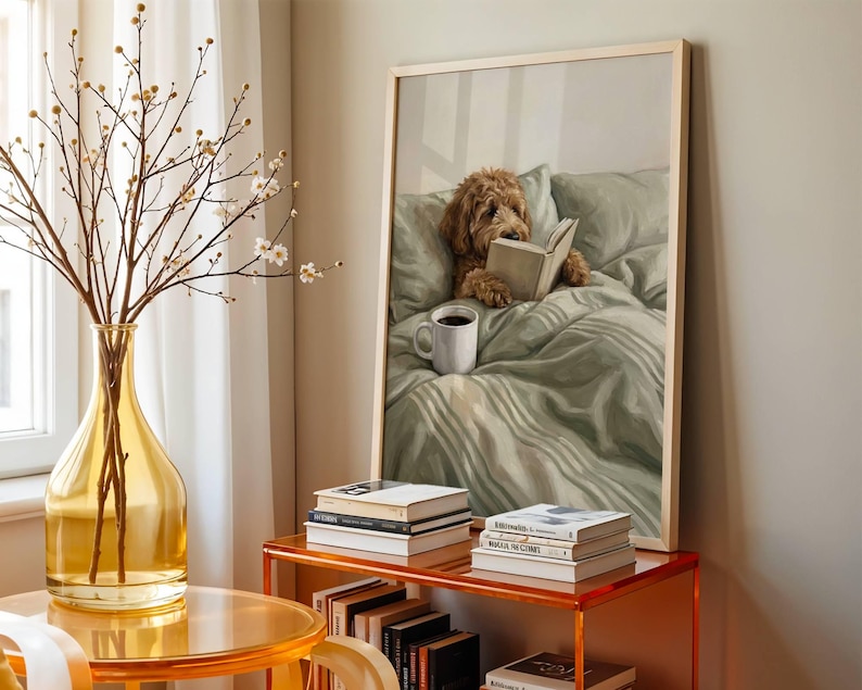 May include: A framed wall art print depicting a golden-brown curly-haired dog lying in a sage green bed, reading an open book with a white mug of coffee nearby. The artwork is displayed on a shelf alongside stacks of books and a glass vase.