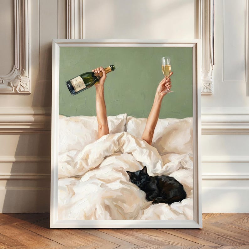 May include: A framed wall art print depicting two arms raised from white bed sheets, one holding a green glass champagne bottle and the other a filled champagne flute. A black cat is curled up sleeping on the white bedding against a sage green wall.