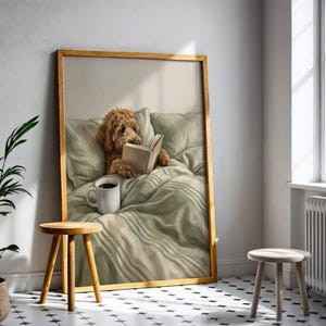 May include: A framed wall art print featuring a golden-brown dog resting in a bed with light green bedding, holding an open book with its paws. A white ceramic mug sits on the rumpled sheets next to the dog, all captured in a soft, painterly style.