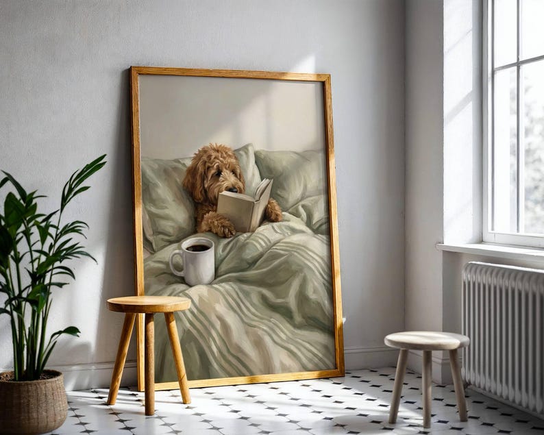 May include: A framed wall art print featuring a golden-brown dog resting in a bed with light green bedding, holding an open book with its paws. A white ceramic mug sits on the rumpled sheets next to the dog, all captured in a soft, painterly style.