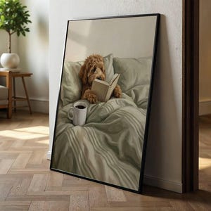 May include: A framed vertical wall art print featuring a golden-brown doodle dog sitting upright in a bed with sage green bedding. The dog is holding an open book with its paws, and a white ceramic mug sits on the rumpled sheets in the foreground.