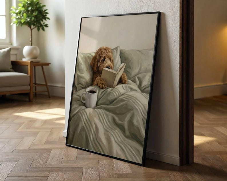 May include: A framed vertical wall art print featuring a golden-brown doodle dog sitting upright in a bed with sage green bedding. The dog is holding an open book with its paws, and a white ceramic mug sits on the rumpled sheets in the foreground.
