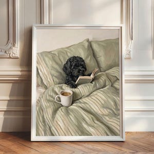 Op de afbeelding: Een ingelijst schilderij van een kleine zwarte hond met krullend haar die rechtop in bed zit met saliegroen gestreept beddengoed. De hond houdt een open boek vast met de voorpoten en er staat een witte keramische mok met koffie op de deken.