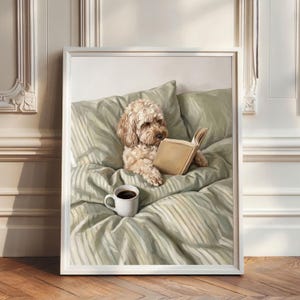 Può includere: Una stampa incorniciata raffigura un cane marrone chiaro che legge un libro su un letto con biancheria da letto verde a righe. Una tazza bianca di caffè è accanto al cane. L'opera d'arte è in una cornice bianca.