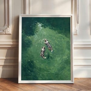 May include: A framed wall art print featuring an overhead view of two blue merle Australian Shepherd dogs swimming in deep green water. The dogs are positioned in the centre of the frame, surrounded by ripples and light reflections on the water surface.
