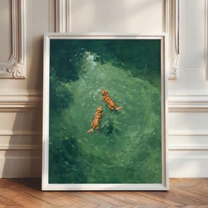 May include: A framed wall art print featuring an oil painting style depiction of two golden-colored dogs swimming in deep green water. The view is from directly above, showing the dogs moving through the rippling, textured surface of the water.