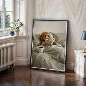 May include: A framed wall art print depicting a golden-brown curly-haired dog sitting upright in a bed with sage green bedding. The dog is holding an open book with its paws, and a white ceramic mug sits on the bed covers in front of the dog.