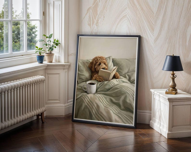 May include: A framed wall art print depicting a golden-brown curly-haired dog sitting upright in a bed with sage green bedding. The dog is holding an open book with its paws, and a white ceramic mug sits on the bed covers in front of the dog.