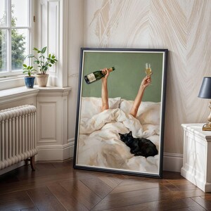 May include: A framed wall art print depicting two arms raised from under white bed sheets, one hand holding a green glass bottle and the other a champagne flute, with a black cat resting on the bedding against a solid sage green background.