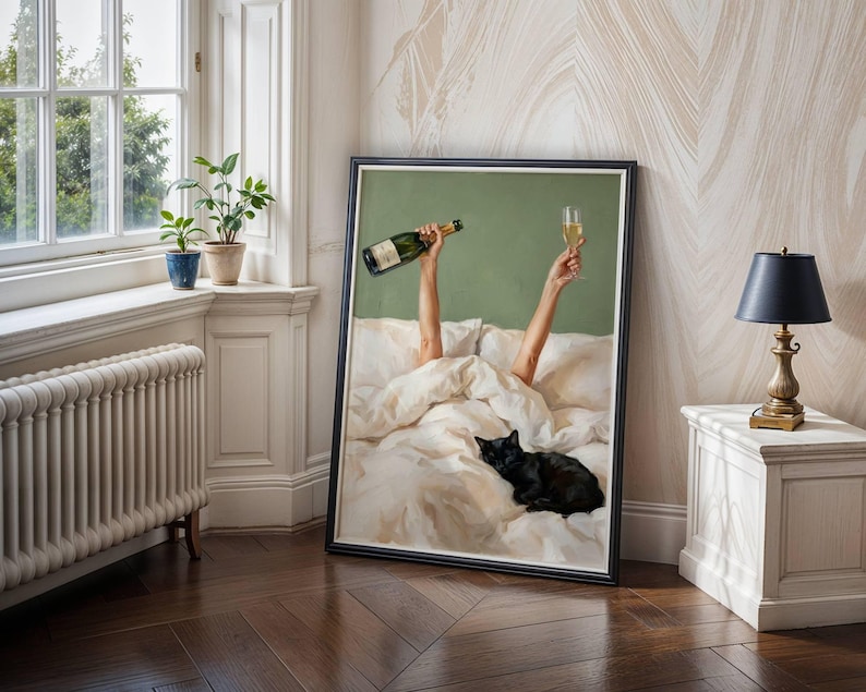 May include: A framed wall art print depicting two arms raised from under white bed sheets, one hand holding a green glass bottle and the other a champagne flute, with a black cat resting on the bedding against a solid sage green background.