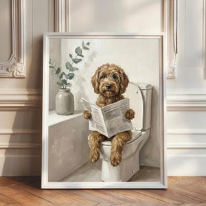 Op de afbeelding: Een ingelijste kunstdruk van een bruine, krulharige hond met een ronde bril met dunne monturen. De hond zit op een wit toilet en leest een krant. Naast het toilet staat een grijze vaas met een groene eucalyptusplant.
