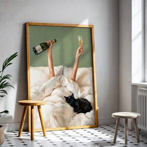 May include: A framed wall art print featuring a green background with two arms reaching up from white bedding, one holding a champagne bottle and the other a champagne flute. A black cat is curled up on the white bed sheets in the foreground.
