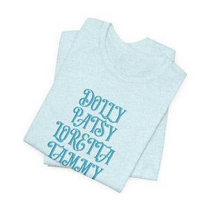 Puede incluir: Camiseta azul claro con los nombres Dolly, Patsy, Loretta y Tammy bordados en una fuente de escritura azul claro. La camiseta está doblada y parece estar hecha de una tela suave y jaspeada. El diseño está centrado en la parte delantera de la camiseta.