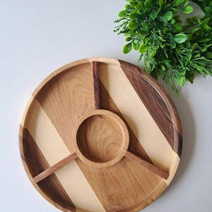 May include: A round wooden serving tray with a central dip bowl and three pie-shaped sections. The tray is made of light and dark wood, with a diagonal striped pattern. A green wreath is in the background.