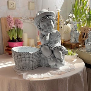 May include: A gray concrete statue of a girl wearing a hat and dress, seated next to a woven basket planter. The statue is on a table with a pink tablecloth, with pink hyacinth flowers in the background.