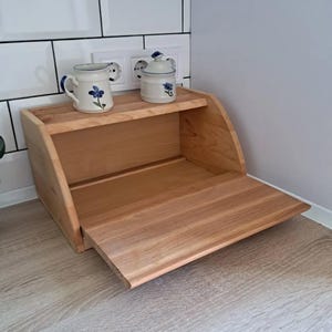May include: A light-colored wooden bread box with a hinged front door. The top shelf holds a small white pitcher and a matching sugar bowl, both decorated with blue floral designs. The bread box is on a wooden surface.