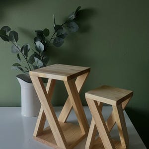 May include: Two wooden side tables with an X-shaped base and square tops. The tables are made of light-colored wood and are different sizes. A white pitcher with greenery is in the background.