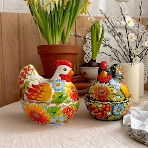Puede incluir: Dos recipientes decorativos de cerámica con forma de gallina. Uno es blanco con diseños florales rojos, amarillos y azules. El otro es negro con girasoles y bayas rojas. Ambos tienen crestas y picos rojos.