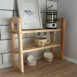 Puede incluir: Una estantería de madera de dos niveles con una impresión enmarcada que dice "BON appétit". Una linterna crema y una taza floral se encuentran en el estante superior. El estante inferior contiene dos platos rosas en forma de corazón.