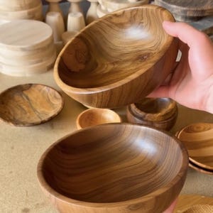 Puede incluir: Cuencos de madera tallados a mano en varios tamaños y formas. Los cuencos son de madera marrón claro con vetas visibles. También se ven otros artículos de madera, como platos pequeños y pedestales.