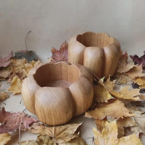 Puede incluir: Dos cuencos de madera tallados con forma de calabaza con un acabado natural de veta de madera marrón claro. Los cuencos presentan un diseño exterior redondeado y segmentado con un centro hueco y una textura suave, sobre hojas de otoño secas.